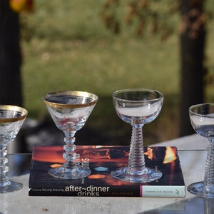 May include: Four clear glass after-dinner drink glasses with gold rims. Two of the glasses have a swirl design on the stem. The glasses are sitting on a book titled "After-Dinner Drinks".