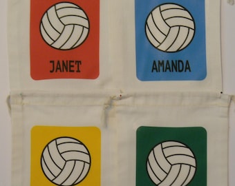 May include: Four white drawstring bags with different colored patches. Each patch features a white volleyball with black lines and the name of a person: Janet, Amanda, Patty, and Bill.