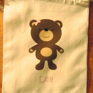 May include: A white drawstring bag featuring a brown bear design. The bear has a pink flower on its head and the name "Lea" is printed below it.