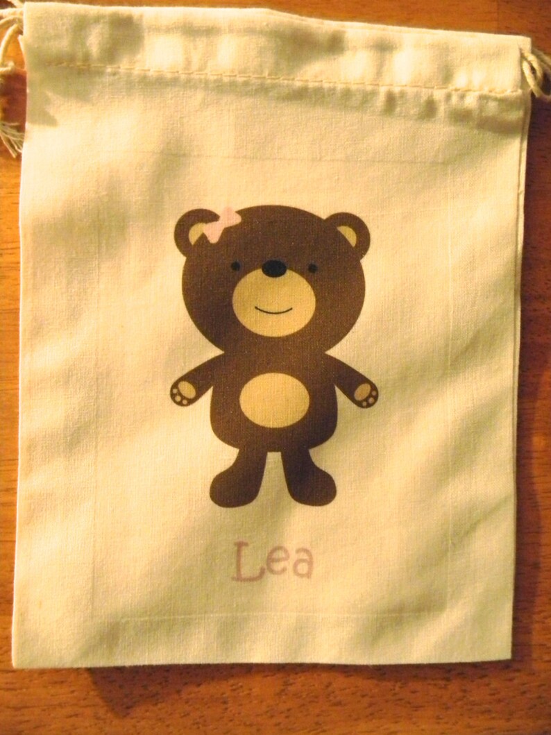 May include: A white drawstring bag featuring a brown bear design. The bear has a pink flower on its head and the name "Lea" is printed below it.
