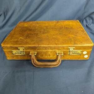 May include: A rectangular brown leather briefcase featuring a weathered, textured surface, a sturdy top handle, and two metallic combination locks on the front. The case has visible signs of wear and two small white circular stickers on the right side.