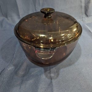 May include: A round, translucent brown glass casserole dish with a matching lid. The dish features two integrated side handles and a central knob on the lid. The glass has a smooth, reflective surface and a deep amber tint throughout the entire piece.