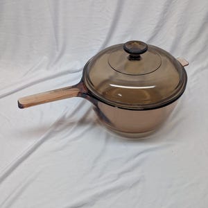 May include: A translucent amber-colored glass saucepan with a matching circular lid. The cookware features a long, integrated handle extending from one side and a small, round knob centered on the top of the lid for easy removal and handling.