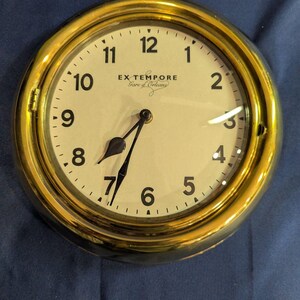 May include: A round wall clock with a polished gold-tone metal frame and a cream-colored face. The dial features black Arabic numerals, black hands, and the text EX TEMPORE Gare d'Orleans printed in black in the center of the clock face.