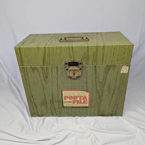 May include: A rectangular olive green storage box with a faux wood grain finish, featuring a brass-colored metal handle on top and a central locking latch. A rectangular label on the front reads "PORTA FILE" in red text with additional details.