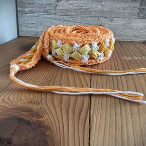 May include: A rolled-up, hand-crocheted belt in shades of orange, yellow, and white. The belt features a floral pattern and long, braided ties. The item is displayed on a wooden surface.