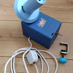 May include: A blue yarn winder with a Union Jack sticker, a white power cord, and a metal clamp with a blue knob. The winder has a cylindrical top and a boxy base. The winder is designed to wind yarn into a ball.