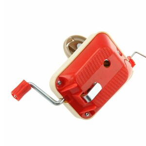 May include: A vintage yarn winder in red and cream. The winder has a red handle and a cream-colored body with a textured red top. The metal crank and other metal parts are visible. The winder is designed to wind yarn into a ball.