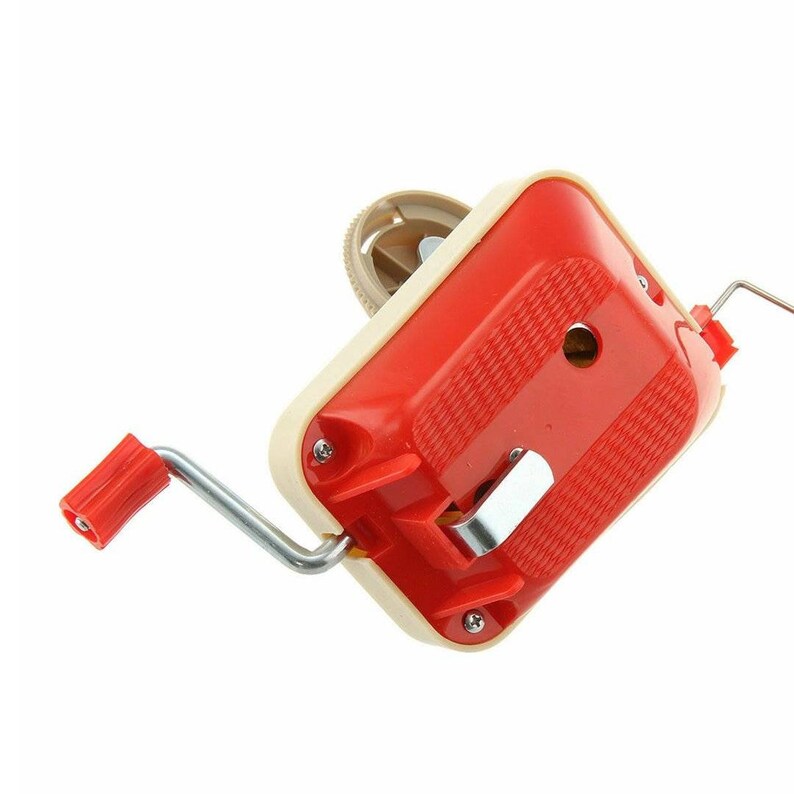 May include: A vintage yarn winder in red and cream. The winder has a red handle and a cream-colored body with a textured red top. The metal crank and other metal parts are visible. The winder is designed to wind yarn into a ball.