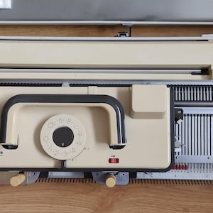 May include: A beige knitting machine featuring a central carriage with a black handle and a circular tension dial numbered zero to ten. The machine includes a long metal needle bed with numerous vertical slots and a top control panel with adjustment knobs.