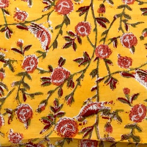 May include: Close-up of a yellow fabric with a floral pattern. The design features red and white flowers, green leaves, and brown stems. The fabric has a repeating pattern of flowers and birds.