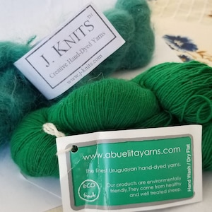 May include: Two skeins of green yarn, one is a fuzzy mohair blend and the other is a smooth worsted weight yarn. The yarn is labeled "J. KNITS Creative Hand-Dyed Yarns" and "www.abuelitayarns.com The finest Uruguayan hand-dyed yarns. ECO friendly. Our products are environmentally friendly. They come from healthy and well treated sheep. Hand Wash/Dry Flat"