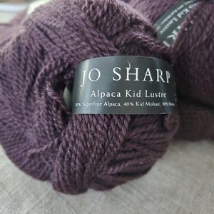 以下が含まれることがあります： 濃い紫色のジョ・シャープ アルパカキッドラスター糸の糸巻き。ラベルには「アルパカキッドラスター」と表示され、糸の組成は、スーパーファインアルパカ30％、キッドモヘア40％、メリノ30％と記載されています。