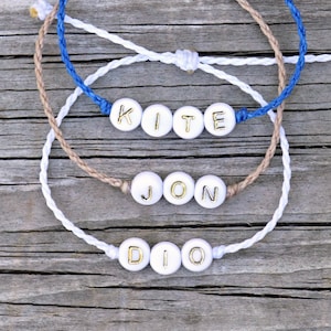 May include: Three friendship bracelets with the names "KITE", "JON", and "DIO" spelled out in gold letters on white beads. The bracelets are made of blue, white, and brown cord.