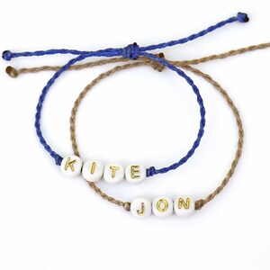 May include: Two friendship bracelets with the names "KITE" and "JON" spelled out in gold letters on white beads. The bracelets are made with blue and brown cord.