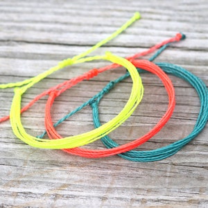 May include: Three colorful friendship bracelets made of woven string. The bracelets are in shades of green, yellow, and orange.