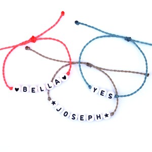 May include: Three friendship bracelets with different colored cords and white beads spelling out the names "Bella", "Yes", and "Joseph".