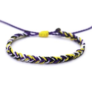 Könnte beinhalten: Geflochtenes Armband mit einem Design der Non-Binary-Pride-Flagge. Das Armband besteht aus gewebten Fäden in Lila, Gelb, Weiß und Schwarz. Es hat verstellbare lila Kordeln und einen gelben Knotenverschluss.