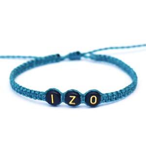 May include: Teal macrame bracelet with black and gold letter beads spelling "IZO". The adjustable bracelet is displayed on a white background. A handmade jewelry item.