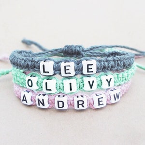 A stack of three bracelets woven from hemp cord with black-and-white plastic letter beads spelling out custom names. The top bracelet is gray, the middle bracelet is a mint light green, and the bottom bracelet is pink.