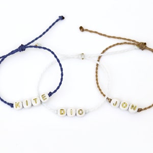 May include: Three friendship bracelets with white beads and gold lettering spelling out the names "KITE", "DIO", and "JON". The bracelets are made with blue, white, and brown cords.