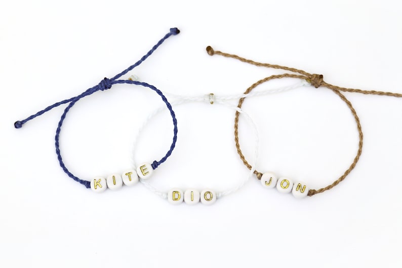 May include: Three friendship bracelets with white beads and gold lettering spelling out the names "KITE", "DIO", and "JON". The bracelets are made with blue, white, and brown cords.