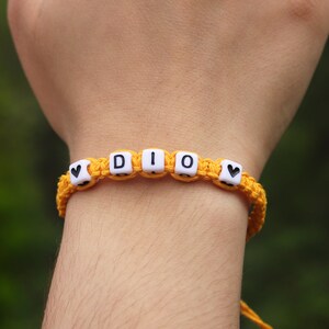 A custom yellow braided hemp bracelet with white letter beads that spell out the word 'DIO' with a black heart bead on each end.