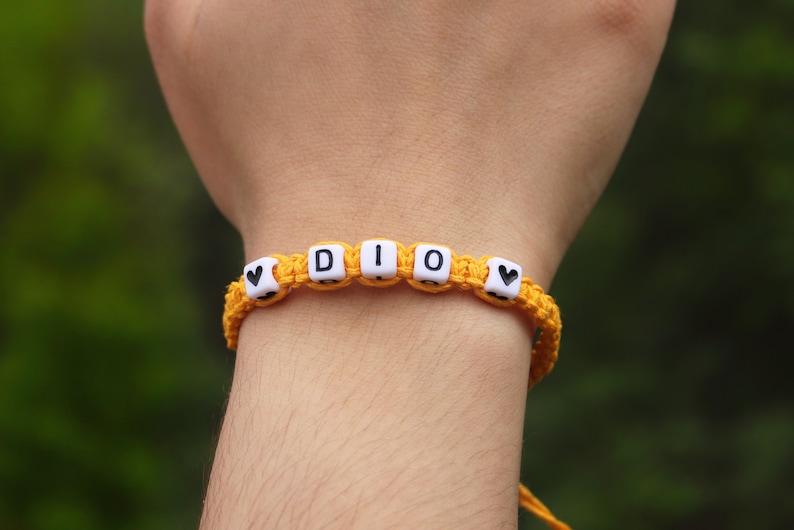 A custom yellow braided hemp bracelet with white letter beads that spell out the word 'DIO' with a black heart bead on each end.