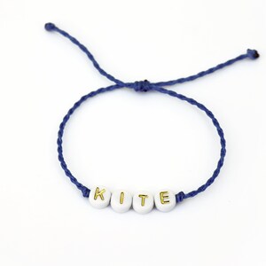 May include: A navy blue string bracelet with white beads that spell out the word "KITE" in gold lettering.
