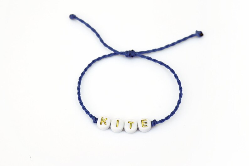 May include: A navy blue string bracelet with white beads that spell out the word "KITE" in gold lettering.