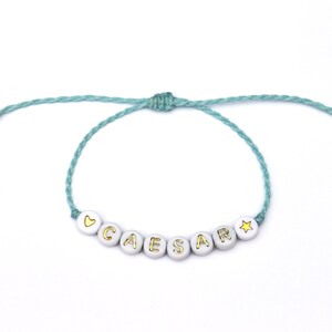 May include: A light blue string bracelet with white beads that spell out the name "CAESAR" with a gold heart and star charm.