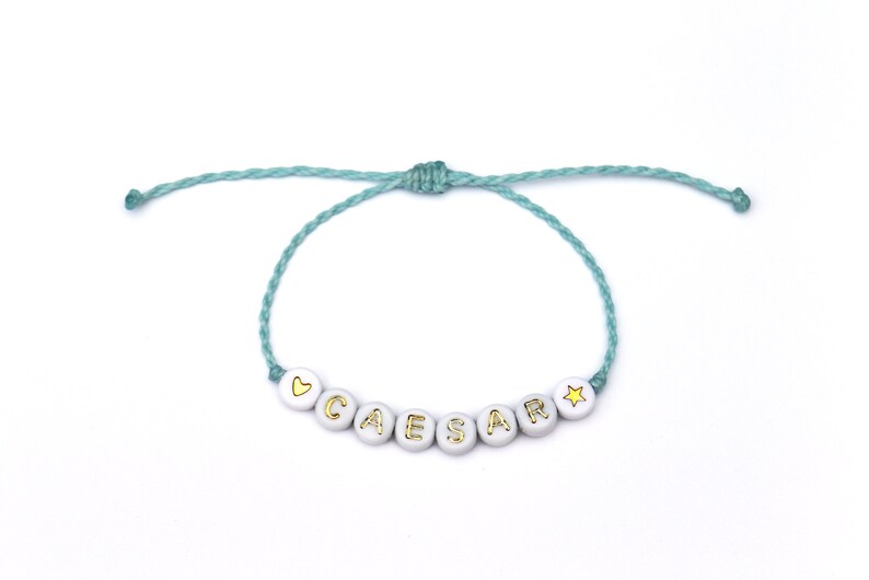 May include: A light blue string bracelet with white beads that spell out the name "CAESAR" with a gold heart and star charm.