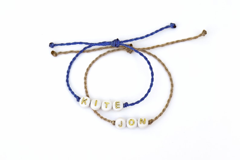 May include: Two friendship bracelets with the names "KITE" and "JON" spelled out in gold letters on white beads. The bracelets are made with blue and brown cord.