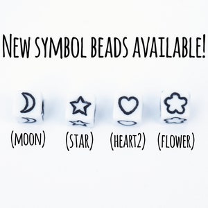 Four white cube beads with black symbols. The symbols are a crescent moon, a star, a heart, and a flower. The text below the beads reads '(MOON) (STAR) (HEART2) (FLOWER)'. NEW SYMBOL BEADS AVAILABLE! is written above the beads.