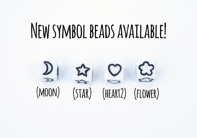 Four white cube beads with black symbols. The symbols are a crescent moon, a star, a heart, and a flower. The text below the beads reads '(MOON) (STAR) (HEART2) (FLOWER)'. NEW SYMBOL BEADS AVAILABLE! is written above the beads.