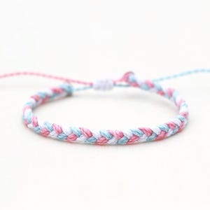 May include: Braided bracelet in pink, white, and light blue. The adjustable bracelet is made of woven cord and features a sliding knot closure. The bracelet is shown on a white background.