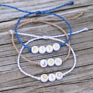 May include: Three friendship bracelets with white beads and gold lettering spelling out the names "KITE", "JON", and "DIO". The bracelets are made with blue, brown, and white string.