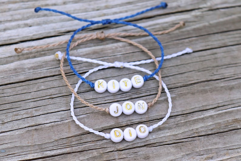 May include: Three friendship bracelets with white beads and gold lettering spelling out the names "KITE", "JON", and "DIO". The bracelets are made with blue, brown, and white string.