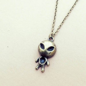 May include: A bronze necklace with an alien pendant. The pendant is a small, cartoon-like alien with large eyes and a simple body.