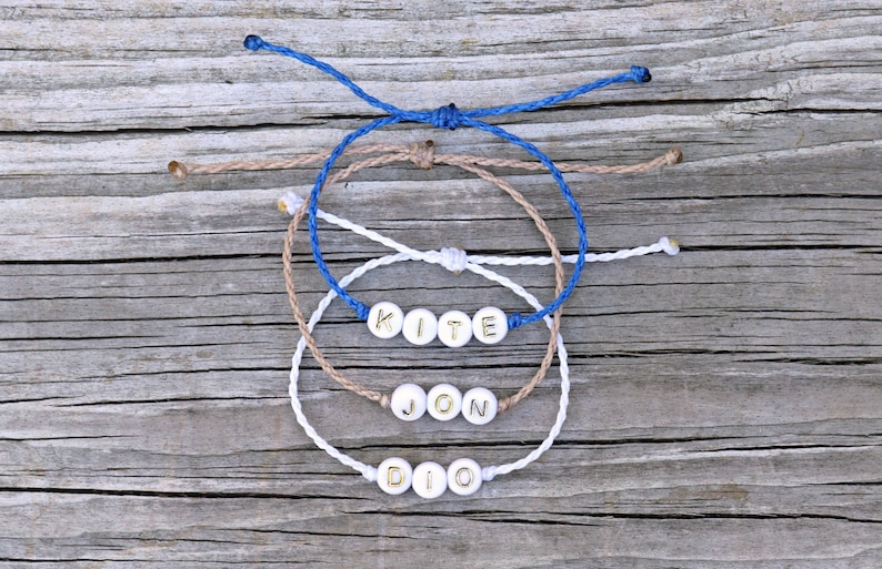 May include: Three friendship bracelets with the names "KITE", "JON", and "DIO" spelled out in gold letters on white beads. The bracelets are made of blue, white, and brown cord.