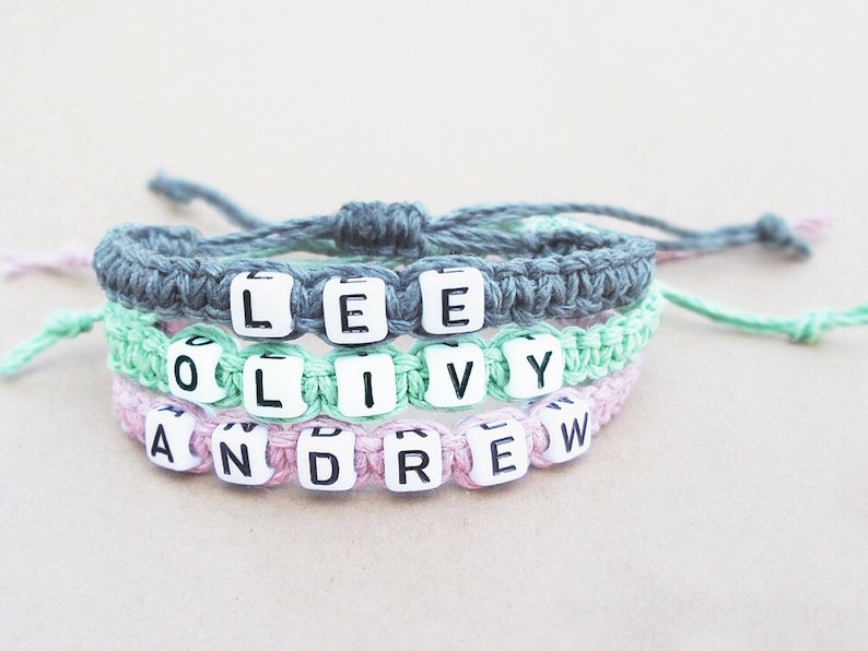 A stack of three bracelets woven from hemp cord with black-and-white plastic letter beads spelling out custom names. The top bracelet is gray, the middle bracelet is a mint light green, and the bottom bracelet is pink.