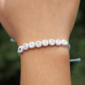 May include: A white beaded bracelet with the name "CAESAR" spelled out in gold letters. The bracelet has a blue string and a heart charm at the beginning and a star charm at the end.