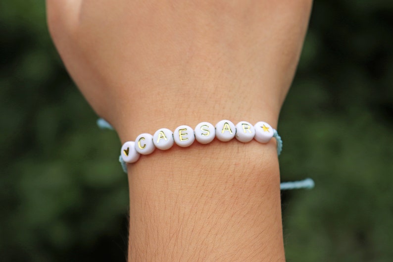 May include: A white beaded bracelet with the name "CAESAR" spelled out in gold letters. The bracelet has a blue string and a heart charm at the beginning and a star charm at the end.