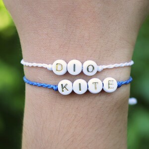 May include: Two friendship bracelets with white beads and gold lettering. The top bracelet spells "DIO" and the bottom bracelet spells "KITE".