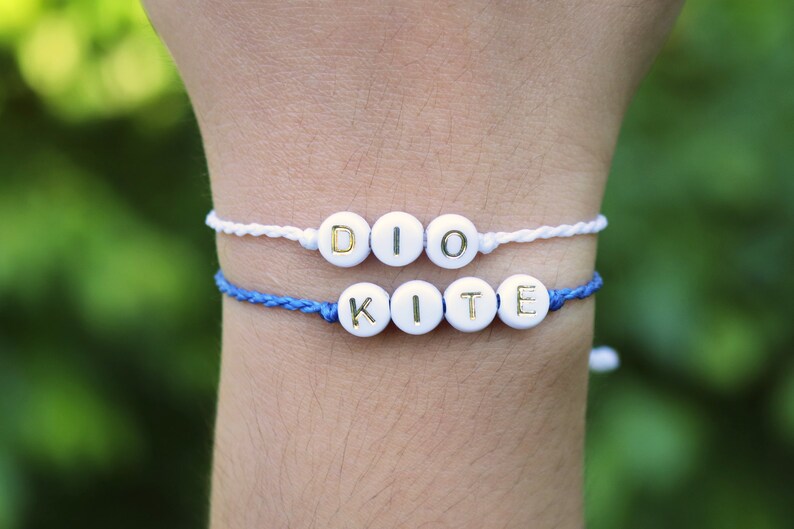 May include: Two friendship bracelets with white beads and gold lettering. The top bracelet spells "DIO" and the bottom bracelet spells "KITE".