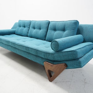 May include: A teal blue mid-century modern sofa with tufted cushions and a matching bolster pillow. The sofa has wooden legs with a unique geometric design.