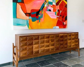 May include: A mid-century modern walnut wood sideboard with a geometric pattern on the doors. The sideboard has four legs and is sitting on a tiled floor. A colourful abstract painting hangs on the wall behind the sideboard.