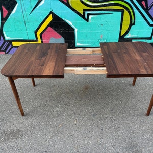 May include: A rectangular, dark brown wooden dining table with rounded corners and four angled legs. The table has an expandable leaf mechanism, with the center section partially extended. The background features a colorful graffiti mural.