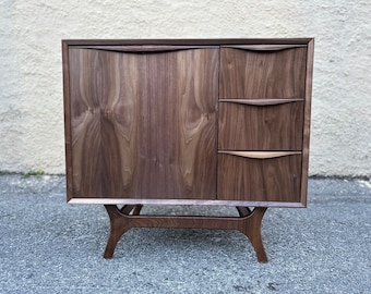 May include: A mid-century modern style wooden cabinet with a dark brown finish. The cabinet has a single door on the left and two drawers on the right. The cabinet is supported by a wooden base with angled legs.