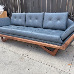 May include: A modern, three-cushion sofa with a blue-grey leather seat and back. The sofa features a wooden frame with angled legs and armrests. The cushions have button detailing.